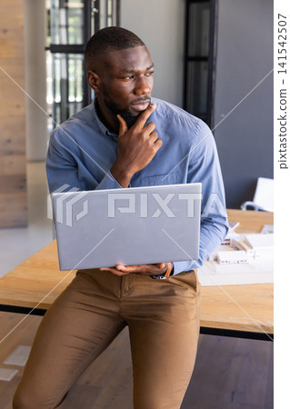 African american man sitting on wood table in modern office holding laptop next to scale mode African american man sitting on wood table in modern office holding laptop next to scale mode 141542507