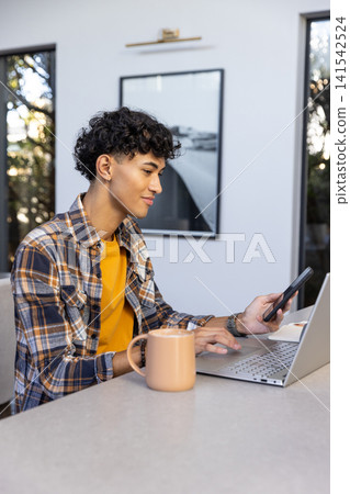 Man sitting at kitchen countertop wearing shirt typing on laptop holding smartphone with mug Man sitting at kitchen countertop wearing shirt typing on laptop holding smartphone with mug 141542524