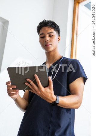 Asian male doctor wearing scrubs, stethoscope checking tablet with smartwatch by window Asian male doctor wearing scrubs, stethoscope checking tablet with smartwatch by window 141542526