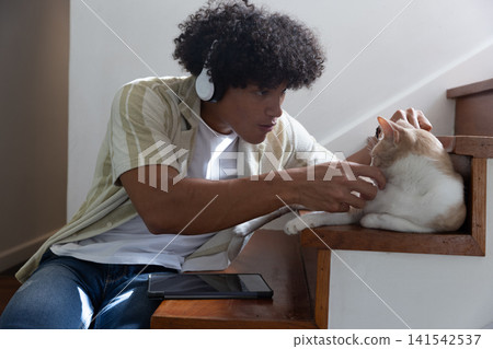African american man on stairs with white headphones petting cat beside tablet at home African american man on stairs with white headphones petting cat beside tablet at home 141542537