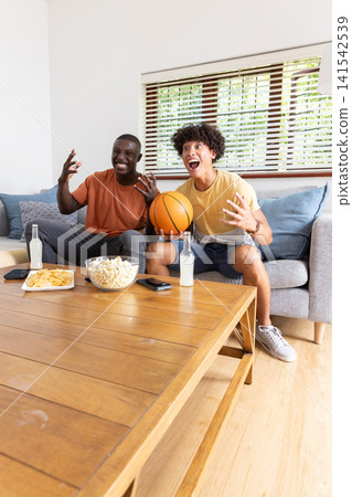 Diverse male friends holding basketball, reacting to game on couch in living room with popcorn Diverse male friends holding basketball, reacting to game on couch in living room with popcorn 141542539