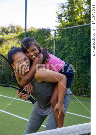 Asian mother and daughter in sportswear giving piggyback ride on tennis court while holding racket Asian mother and daughter in sportswear giving piggyback ride on tennis court while holding racket 141542551
