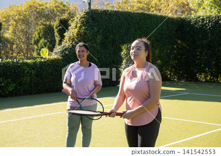 Two female friends in sportswear standing on green tennis court, holding rackets Two female friends in sportswear standing on green tennis court, holding rackets 141542553