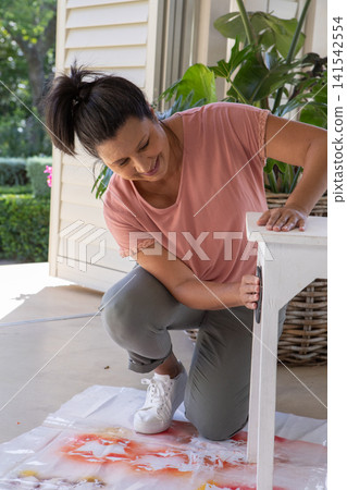 Senior woman kneeling on paint-stained drop cloth sanding small wooden side table on porch Senior woman kneeling on paint-stained drop cloth sanding small wooden side table on porch 141542554