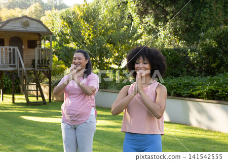Hispanic mother and african american daughter practicing yoga pose in garden with wooden playhouse Hispanic mother and african american daughter practicing yoga pose in garden with wooden playhouse 141542555