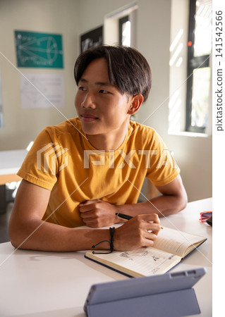 Asian teenage boy sitting at white desk in classroom studying with tablet and notebook Asian teenage boy sitting at white desk in classroom studying with tablet and notebook 141542566