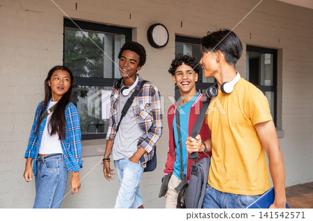 Diverse student friends walking and talking along campus corridor with backpacks and tablet 141542571