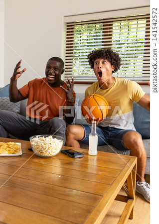 Diverse male friends sitting on sofa in living room holding basketball, grabbing popcorn Diverse male friends sitting on sofa in living room holding basketball, grabbing popcorn 141542572