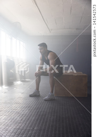 Man in twenties resting on box in gym beside weight plates, catching breath between training sets Man in twenties resting on box in gym beside weight plates, catching breath between training sets 141542573