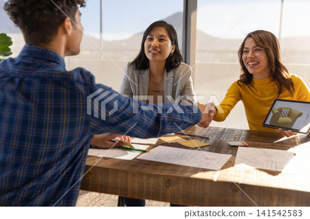 Business associates shaking hands in office showing couch on tablet amid blueprints and sketches 141542583