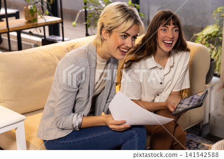 Female colleagues leaning forward reviewing printed documents and tablet on sofa in office Female colleagues leaning forward reviewing printed documents and tablet on sofa in office 141542588