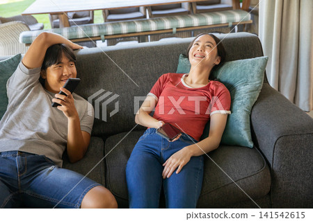 Asian friends reclining on sofa with teal cushion at home using smartphones and smiling Asian friends reclining on sofa with teal cushion at home using smartphones and smiling 141542615