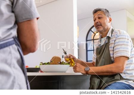 Couple preparing fresh salad at kitchen island, pouring olive oil into lettuce and tomato Couple preparing fresh salad at kitchen island, pouring olive oil into lettuce and tomato 141542616