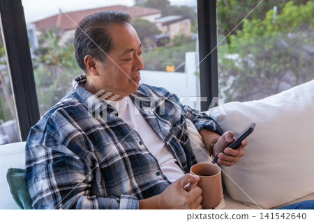 Asian man sitting on sofa near window holding smartphone and mug in living room Asian man sitting on sofa near window holding smartphone and mug in living room 141542640