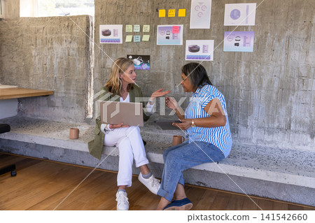Diverse female coworkers brainstorming on bench in office lounge with laptop, sticky notes Diverse female coworkers brainstorming on bench in office lounge with laptop, sticky notes 141542660