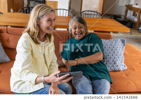 Senior female friends holding virtual reality headset and laughing on sofa at home Senior female friends holding virtual reality headset and laughing on sofa at home 141542666