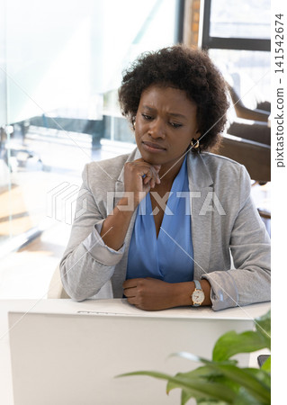African american woman sitting at desk in modern office frowning at laptop screen African american woman sitting at desk in modern office frowning at laptop screen 141542674