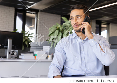 Man holding smartphone to ear sitting at office desk with monitors, copy space Man holding smartphone to ear sitting at office desk with monitors, copy space 141542685