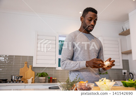 African american man peeling garlic at kitchen counter with cutting board, spice jars African american man peeling garlic at kitchen counter with cutting board, spice jars 141542690