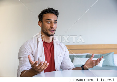 Man in twenties sitting at guest room table speaking to camera with pillows on headboard 141542691