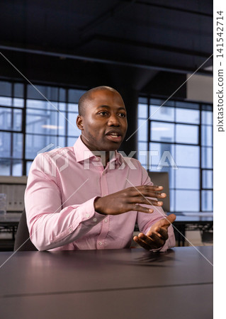 African american man gesturing while sitting at conference table in office with beams, windows African american man gesturing while sitting at conference table in office with beams, windows 141542714