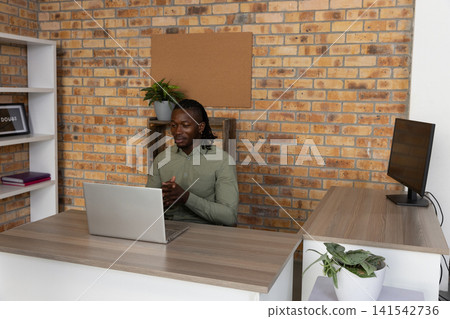African american man sitting at desk in home office in light green shirt gesturing at laptop African american man sitting at desk in home office in light green shirt gesturing at laptop 141542736