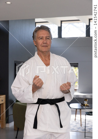 Mature male standing in living room wearing white gi and black belt holding clenched fists Mature male standing in living room wearing white gi and black belt holding clenched fists 141542741