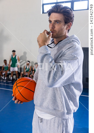Coach and diverse teenage team coaching on court by bench holding basketball blowing whistle Coach and diverse teenage team coaching on court by bench holding basketball blowing whistle 141542749