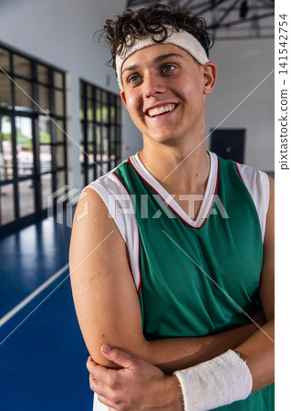 Teenage male basketball player in green jersey standing with crossed arms on court near windows Teenage male basketball player in green jersey standing with crossed arms on court near windows 141542754