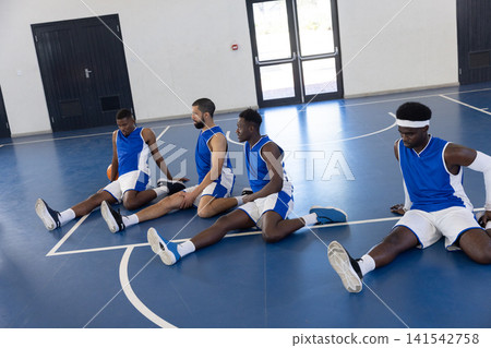 Diverse male teammates stretching legs on gym court in sportswear with basketball, court lines Diverse male teammates stretching legs on gym court in sportswear with basketball, court lines 141542758