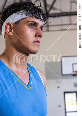 Teen male basketball player standing near gym wall wearing jersey, headband by hoop, copy space Teen male basketball player standing near gym wall wearing jersey, headband by hoop, copy space 141542762