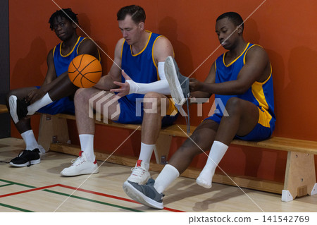 Diverse male basketball teammates sitting on bench in gym holding orange basketball Diverse male basketball teammates sitting on bench in gym holding orange basketball 141542769