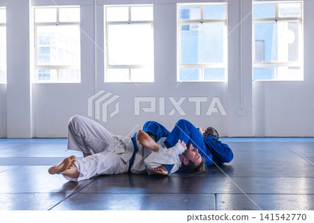 Diverse male training partners practicing grappling on dojo mat in gis and belts, copy space Diverse male training partners practicing grappling on dojo mat in gis and belts, copy space 141542770