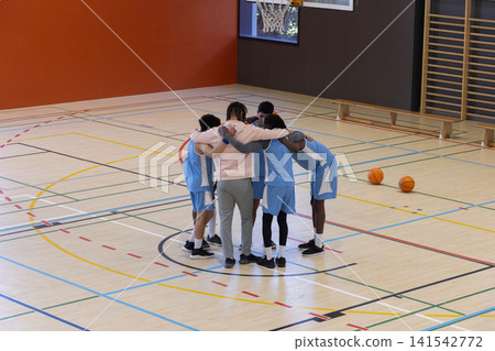 Diverse male basketball team huddling under hoop on polished court with orange basketballs nearby Diverse male basketball team huddling under hoop on polished court with orange basketballs nearby 141542772