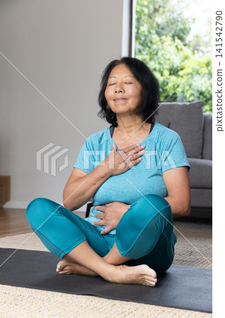 Senior Asian woman wearing sportswear practicing breathing on yoga mat in living room beside sofa Senior Asian woman wearing sportswear practicing breathing on yoga mat in living room beside sofa 141542790