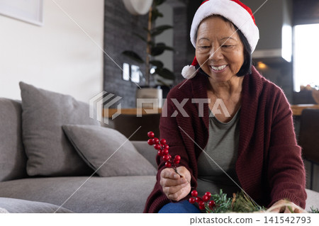 Senior asian woman in red cardigan, santa hat arranging greenery on couch, copy space Senior asian woman in red cardigan, santa hat arranging greenery on couch, copy space 141542793