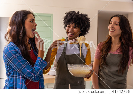 Diverse female friends preparing batter at home kitchen island with glass bowl whisk, aprons Diverse female friends preparing batter at home kitchen island with glass bowl whisk, aprons 141542796