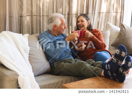Senior couple sitting on sofa holding red mugs beside wooden coffee table in living room Senior couple sitting on sofa holding red mugs beside wooden coffee table in living room 141542811