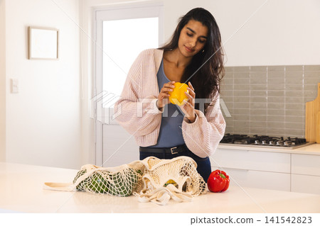 Asian woman holding yellow bell pepper and inspecting mesh bags and red pepper on island countertop Asian woman holding yellow bell pepper and inspecting mesh bags and red pepper on island countertop 141542823