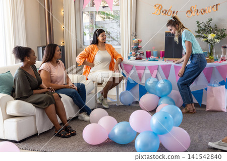 Diverse female friends celebrating pastel baby shower in living room with balloons, cupcakes Diverse female friends celebrating pastel baby shower in living room with balloons, cupcakes 141542840