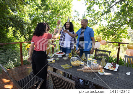 Diverse friends raising champagne flutes around deck table with balloons, hats, bunting, cake Diverse friends raising champagne flutes around deck table with balloons, hats, bunting, cake 141542842