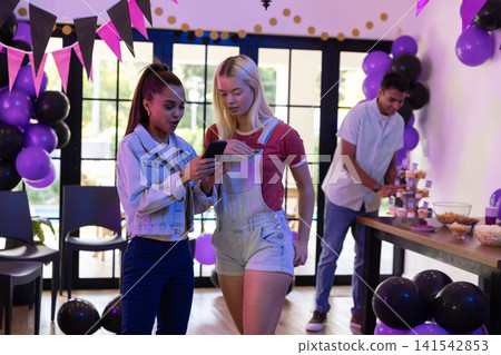 Diverse female friends examining smartphone at festive living room party with balloons, cupcakes Diverse female friends examining smartphone at festive living room party with balloons, cupcakes 141542853