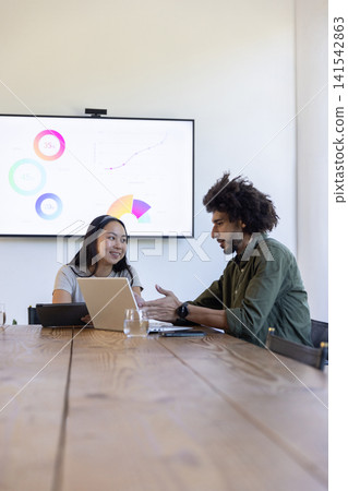 Diverse coworkers in conference room discussing data on display, reviewing laptop at table Diverse coworkers in conference room discussing data on display, reviewing laptop at table 141542863