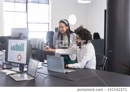 Diverse coworkers reviewing data on large monitor at desk in open-plan office, laptops, tablet 141542871