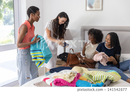 Diverse female friends packing suitcase with clothes and passport in bedroom with lamp and handbag Diverse female friends packing suitcase with clothes and passport in bedroom with lamp and handbag 141542877
