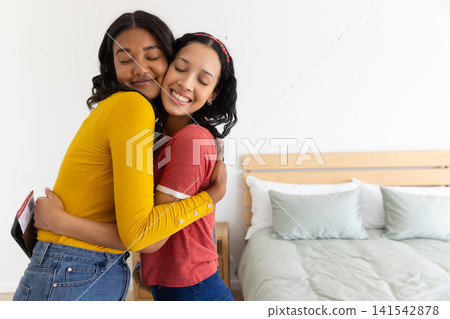 Diverse mom and teenage daughter hugging in bedroom with duvet pillows on wooden bedframe Diverse mom and teenage daughter hugging in bedroom with duvet pillows on wooden bedframe 141542878