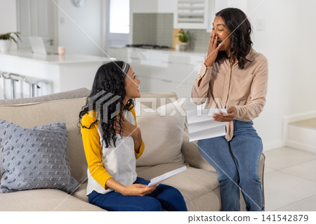 Diverse mom and teenage daughter reacting while reading letters on beige couch by kitchen island 141542879