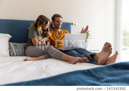 Couple working on laptop sitting on bed in bedroom and holding pen near potted plant 141542892