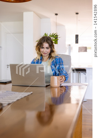 Woman working on laptop and holding coffee mug at home kitchen table with smartphone and papers Woman working on laptop and holding coffee mug at home kitchen table with smartphone and papers 141542896