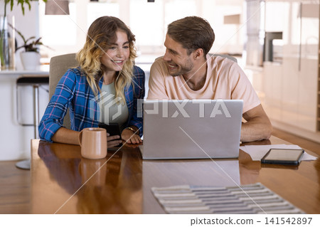 Couple sitting at table in kitchen, holding smartphone and leaning toward laptop beside coffee mug 141542897